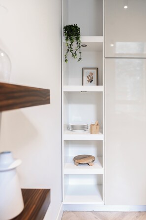 Open shelving with minimalist decor—stacked plates, wooden accents, a trailing plant, and framed art—adds warmth and character to the modern kitchen.