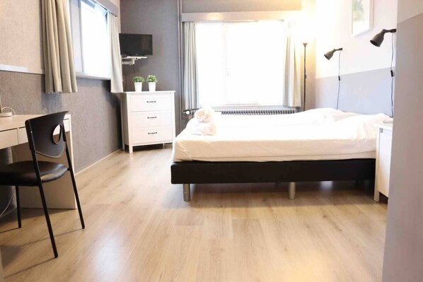 Features a comfortable twin bed, expansive window, bedside lamp, flat-screen TV, and a work table with chair. Perfect for solo travelers or friends seeking a cozy, light-filled space for relaxation and entertainment.