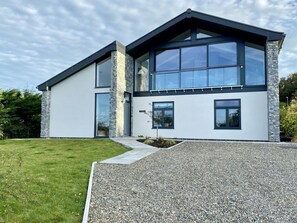 Front of house large windows with balcony, gravelled yard with grass area. paving slabs