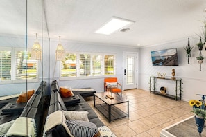 Living room with wall covered in mirrors, large sofa, armchair, side tables, TV, decorations with plants, exterior windows, access door.