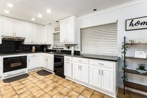A spacious, modern kitchen featuring white cabinetry with sleek handles and dark granite countertops. The kitchen is equipped with an oven, a dishwasher, and a stainless-steel sink under a window with blinds. The backsplash is a glossy black, adding a stylish contrast to the white cabinets. The tiled floor complements the warm and inviting feel of the space. On the right, there...