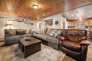 Living Room with  Fire Place - Warm wood ceilings glow in the living room, furnished with a plush sectional and armchair. Settle in and watch a film on the 50-in smart TV while the wood-burning fireplace crackles. Features a DVD collection and board game selection!