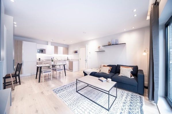 Open-plan living room with a fully equipped kitchen, sofa bed suitable for the 5th and 6th guests,  a dedicated desk with a 27-inch monitor, and a smart TV.