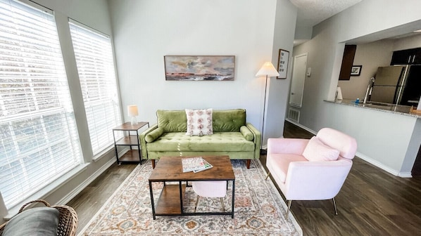Bright and inviting living room featuring a green couch, pink single sofa, and a cozy layout with a peek into the kitchen.