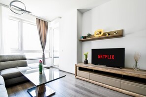 Step into your sleek, urban retreat after a bustling day in Sofia's heart. This bright, contemporary living space, framed by large windows inviting in natural light, offers a serene spot to unwind. A plush, sectional sofa beckons you to relax while the smart TV—currently displaying Netflix—promises entertainment at your fingertips. With elegant, minimalist décor and touches of ...