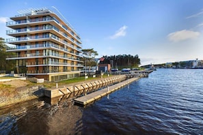 A modern residential building by the water with a pier in the foreground and a blue sky in the background.