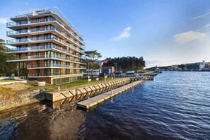 A modern residential building by the water with a pier in the foreground and a blue sky in the background.