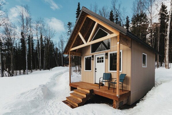 Tiny cabins exterior featuring a charming wooden wall, inviting white door, and two cozy chairs for enjoying the surrounding beauty.