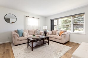 Our expansive living room features a cozy beige corner sofa, adorned with large windows dressed in sheer curtains, accompanied by a dark wooden coffee table, crafting a luminous and welcoming ambiance.