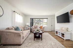 Our bright modern living room is illuminated by sunlight, boasting a comfortable sectional, stylish TV arrangement, and inviting love seat by the window, all unified by a trendy white rug. This is a space exuding contemporary coziness.