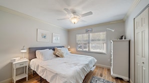 A small bedroom with a bed, two nightstands, a dresser, and a ceiling fan. The room has light-colored walls, wood flooring, a window with blinds, and framed artwork on the walls.