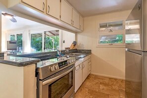 A modern kitchen with bright white cabinetry, stainless steel appliances, and a view of lush greenery through a large window, bathing the space in natural light