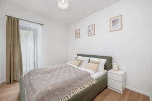 A bedroom with a large bed covered in a beige throw. The room is simply decorated with light curtains and framed pictures on the walls.