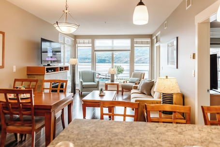 Experience serene lake and mountain views from this cozy living room with a beige sofa, armchairs, and a stylish dining area for six. Warm wood accents and neutral tones create a relaxing retreat.