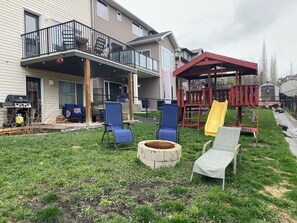 Backyard features patio with BBQ and furniture; play structure; fire pit