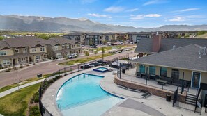 A residential complex featuring a swimming pool, hot tub, and lounge area. The complex consists of two-story townhouses and multi-story buildings, with mountains visible in the distance under a clear sky.