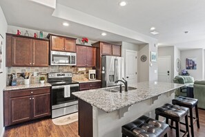 Modern kitchen with dark wooden cabinets, stainless steel appliances, granite countertops, and a large island with three leather stools. Open layout with a living area in the background.