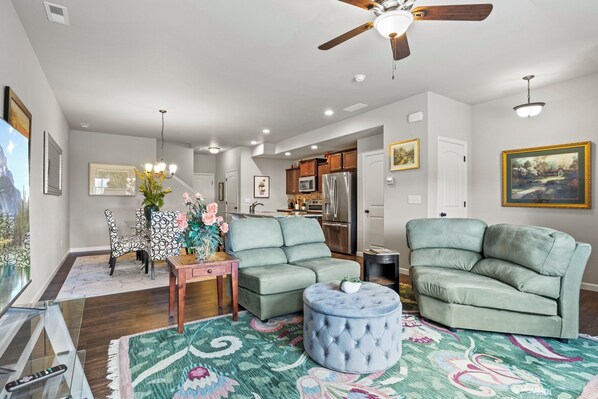 Modern living room with light gray walls and dark hardwood floors, furnished with green seating, a floral-patterned rug, and various wall art. The dining area and kitchen are visible in the background.