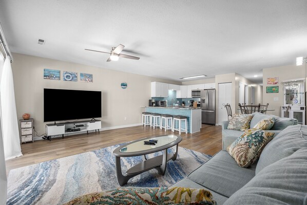 Spacious and inviting living room with a comfortable sofa, vibrant throw pillows, a sleek coffee table, a large flat-screen TV, and modern decor. The room is well-lit with natural light pouring in through the windows, creating a warm atmosphere.