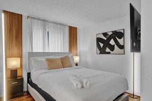 Modern bedroom with a comfortable queen bed, stylish wooden accents, and soft lighting. Features plush pillows, fresh linens, and a large TV for relaxation. Minimalist decor with a touch of elegance. Perfect for a restful stay.