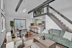 Stylish open-concept living space with high ceilings, modern kitchen, and loft. Features a cozy living area, dining table, and bar seating. Abundant natural light and contemporary decor create a welcoming ambiance. Perfect for relaxation and gatherings.