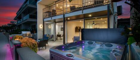 A luxurious two-story beachfront home illuminated at sunset, featuring expansive glass windows, multiple outdoor seating areas, and a shared tub. This stunning property offers a perfect blend of relaxation and sophistication with breathtaking views of the