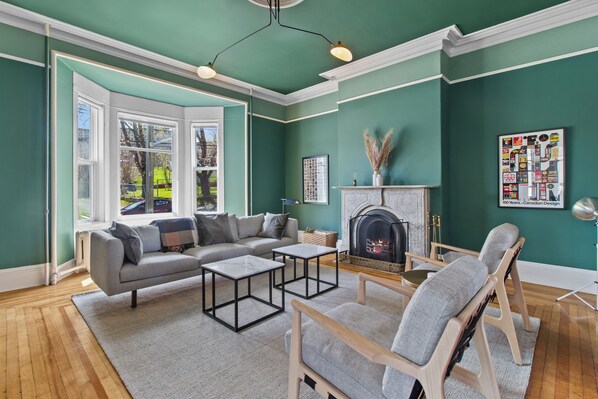 Relax in the beautifully furnished living room, featuring elegant Victorian architecture, rich green walls, and abundant natural light from the bay windows