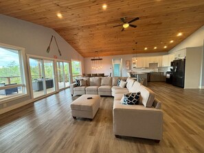 The open-concept living area is spacious and inviting, featuring a comfortable sectional sofa, modern kitchen, and large windows that flood the space with natural light. The wooden ceiling and stylish decor add to the cozy, yet contemporary feel.
