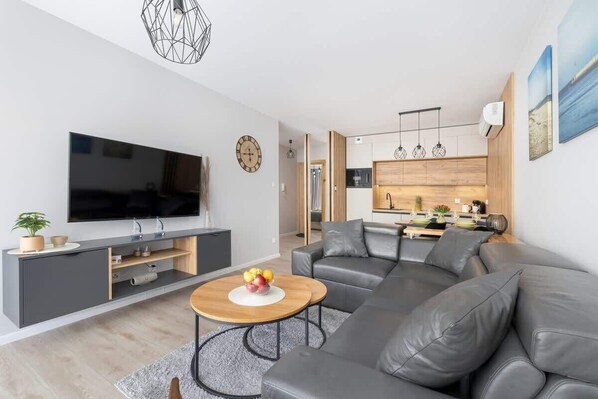 Ekskluzywny salon z aneksem kuchennym. Pomieszczenie jest wyposażone w  sofą narożną, TV i wyjście na balkon.