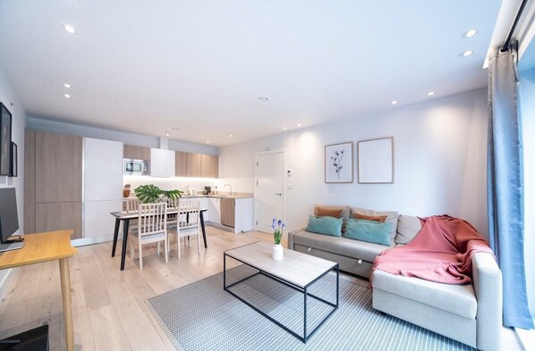 Open-plan living room with a fully equipped kitchen, sofa bed suitable for the 5th and 6th guests,  a dedicated desk with a 27-inch monitor, and a smart TV.