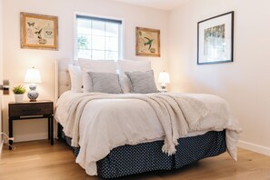 Nestle into the cozy bed with tufted headboard and soft, patterned pillows. The calming blue and white hues create a serene retreat, while framed artwork and natural light enhance the peaceful ambiance.