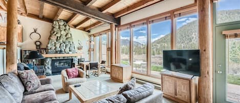 A cozy cabin living room with wooden beams, large windows showcasing a scenic mountain view, a stone fireplace, a sectional sofa, a wooden coffee table, and a TV on a stand.