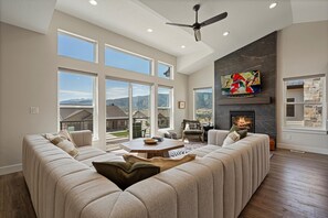 Spacious living room featuring floor-to-ceiling windows, recessed lighting, a large screen TV, a stone fireplace, and a luxurious sectional sofa.