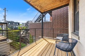 Close view of the property rooftop terrace with a modern metal pergola, wooden decking, a lounge seating area with black chairs and a coffee table, a metal staircase leading up from the ground level, and a lush green wall bordering the terrace.