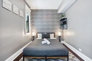 Cozy bedroom with a grey patterned wallpaper wall, a queen sized bed with grey linens and white towels, two bedside tables with lamps, and a window with natural light.