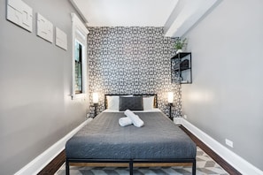 Cozy bedroom with a grey patterned wallpaper wall, a queen sized bed with grey linens and white towels, two bedside tables with lamps, and a window with natural light.
