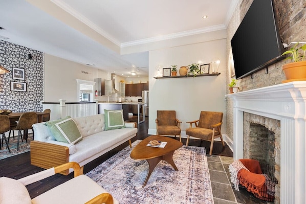 Wide view of the living area with a white sofa, two armchairs, a coffee table, a fireplace, a flat-screen TV, a dining area, and a glimpse of a modern kitchen with stainless steel appliances.