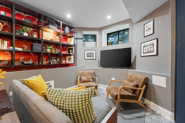 Cozy basement lounge with a grey sofa, two armchairs, a TV, a patterned wallpaper wall, and a black metal bookshelf.
