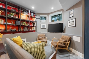 Cozy basement lounge with a grey sofa, two armchairs, a TV, a patterned wallpaper wall, and a black metal bookshelf.