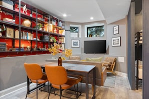 Back view of basement lounge with a grey sofa, two armchairs, a TV, a patterned wallpaper wall, and a black metal bookshelf.