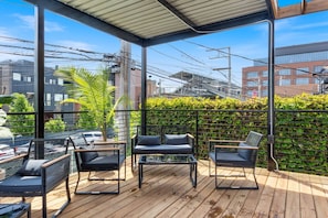 Close view of the property rooftop terrace with a modern metal pergola, wooden decking, a lounge seating area with black chairs and a coffee table, a metal staircase leading up from the ground level, and a lush green wall bordering the terrace.