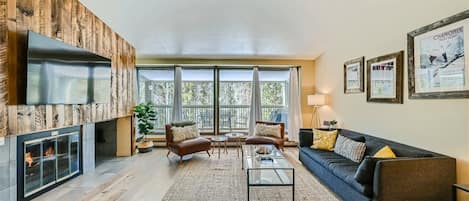 A bright living room with a grey sofa, glass coffee table, two chairs, a fireplace, a large wall-mounted TV, and floor-to-ceiling windows with curtains leading to a balcony.