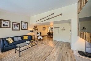 A modern living room with a dark sofa, a glass coffee table, and wall decor. The space features wooden flooring and a dining area visible in the background. Two vintage skis hang on the wall.