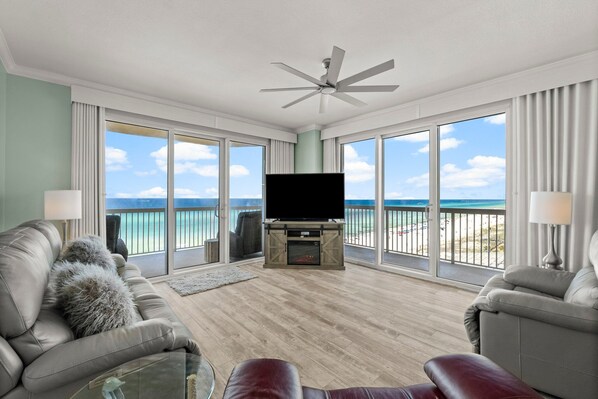 The living room offers comfortable recliners and a reclining couch, large flat screen television, electric fireplace, and floor-to-ceiling corner views of the sugar white sands and emerald green waters.