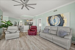 The living room offers comfortable recliners and a reclining couch, large flat screen television, electric fireplace, and floor-to-ceiling corner views of the sugar white sands and emerald green waters.