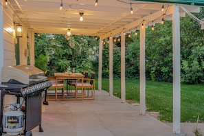 Covered patio gathering area with lights, large table, grass, shade and bbq