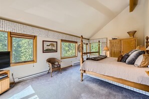 A spacious bedroom with large windows that offer a view of trees, a wooden bed with beige bedding, a wicker chair, a TV, and wooden furniture including a dresser and side tables.