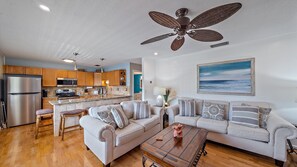 A cozy living room and kitchen area featuring a ceiling fan, beige sofas with patterned cushions, a wooden coffee table, a beach-themed painting, stainless steel appliances, and wooden cabinets.