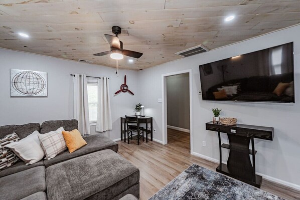 The living room contains plush seating, a flat-screen TV, and a dedicated workspace with ample lighting and a ceiling fan.