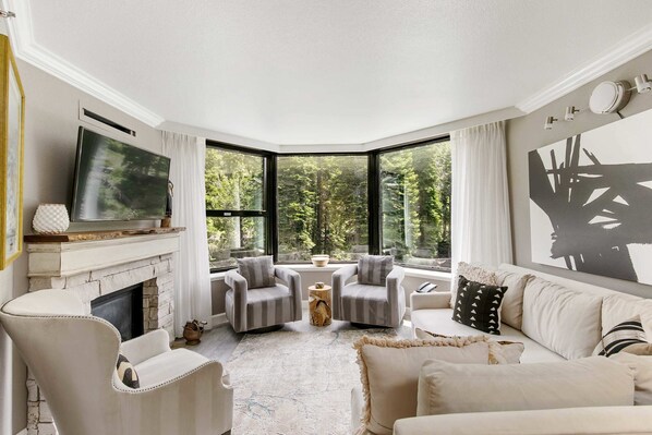 Living Room: L-shaped couch, 3 club chairs, 46” LG TV, gas fireplace, soaring windows framing impressive forested views